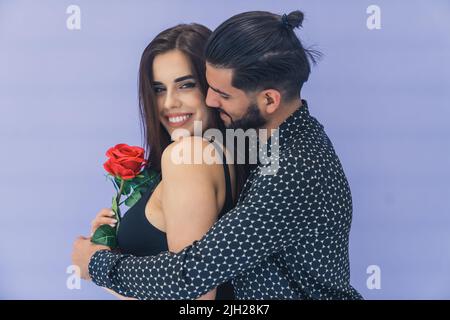 Blumenüberraschung. Dunkelhaariger, bärtiger, hübscher kubanischer Mann umarmt seine geliebte junge Frau von hinten und schenkt ihr eine wunderschöne rote Rose. . Hochwertige Fotos Stockfoto