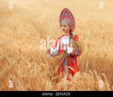 Kleines Mädchen im russischen nationalen Sarafan und ein Kokoschnik, der am Sommertag in einem goldenen Weizenfeld steht Stockfoto