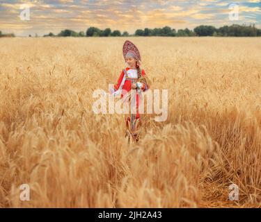 Kleines Mädchen im russischen nationalen Sarafan und ein Kokoschnik, der am Sommertag in einem goldenen Weizenfeld steht Stockfoto