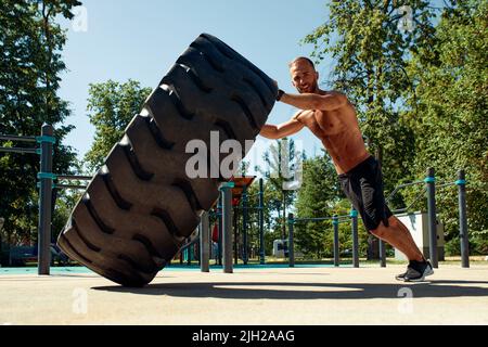 Starker Mann Training Workout Lifting großen Reifen Outdoor-Fitnessraum. Sportlicher junger Erwachsener kaukasischer Kerl Flip großes Rad auf die Natur. Handgefertigte Ausrüstung Sport Stockfoto