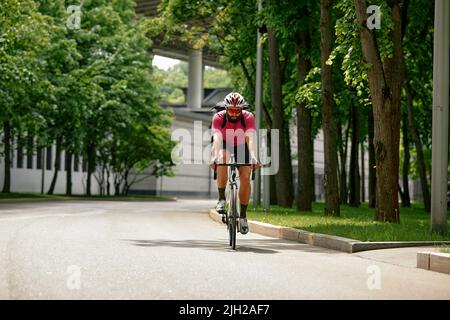 Mann in Sonnenbrillen rast vor dem Hintergrund des Sommerparks entlang der Straße. Mann mit Fahrrad-Hobby fährt im Park. Stockfoto