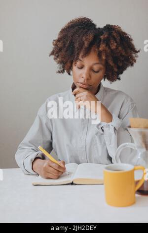 Eine Frau mit lockigen Haaren notiert sich in einem Notizbuch in der Küche. Stockfoto