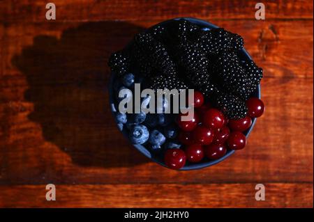 Blaue Keramikschale mit frischen, reifen und verzehrfertigen Bio-Blaubeeren, Brombeeren und Kirschen auf einem hölzernen Hintergrund. Stockfoto