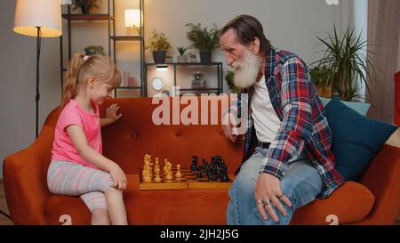 Lächelnder älterer Großvater, der im Wohnzimmer mit einer Enkelin Schach spielt. Im Ruhestand Großeltern genießen Spiel Brettspiel, ruht sich auf der Couch mit Kind Mädchen Kind. Familienaktivitäten Stockfoto