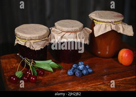 Frische reife Beeren und Früchte auf einem rustikalen Holztisch neben einer Auswahl an hausgemachten Konfitüren und Marmeladen. Stillleben. Stockfoto