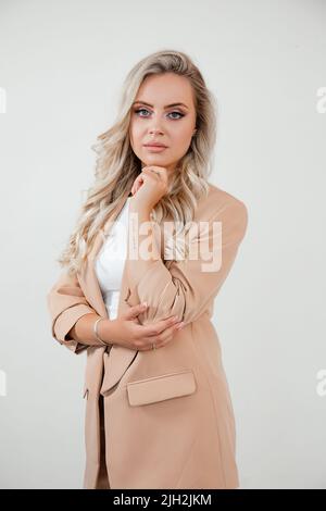 Die stilvolle Business Lady mit langen, gepflegten Locken in der Business Jacket steht auf weißem Hintergrund und blickt in die Kamera. Stockfoto