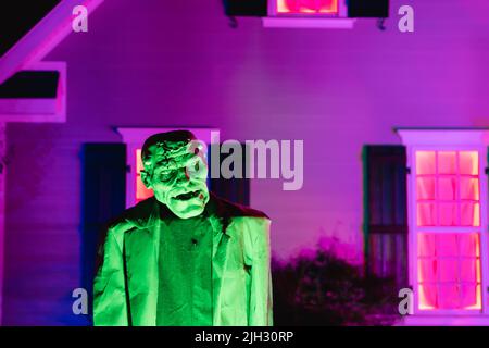 Frankenstein, ein unheimliches, lebensgroßes Monster, das grün vor einem lila beleuchteten Haus im Oktober an Haloween glühte Stockfoto