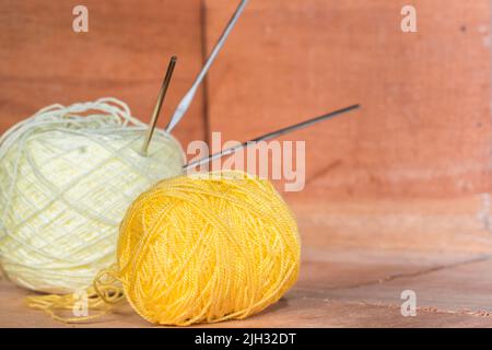 Zwei Rollen gelbes und weißes Garn zum Häkeln, mit Nadeln darauf geklebt, auf einem orangefarbenen Holzhintergrund. Garn bereit zum Häkeln zu Hause. Con Stockfoto