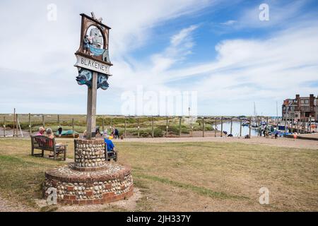 Blakeney Dorfschild abgebildet neben Bänken besetzt von Urlaubern an der Nord-Norfolk-Küste im Juli 2022. Stockfoto