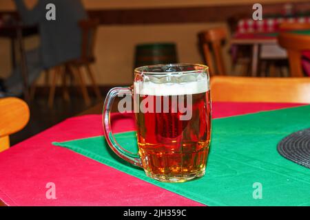 Nahaufnahme eines Glases frischen, schäumenden Bieres auf einem Tisch, der mit einer rot-grünen Tischdecke bedeckt ist. Stockfoto