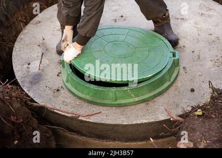 Ein Arbeiter installiert einen Kanalschacht an einem Klärbecken aus Betonringen. Bau von Kanalisationsnetzen für Landhäuser. Stockfoto