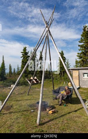 Frau, die im Sommer unter einer Tipi-Struktur in der Nähe der nördlichen indigenen Gemeinschaft Deline, Northwest Territories, Kanada, bei der Feuergrube sitzt Stockfoto