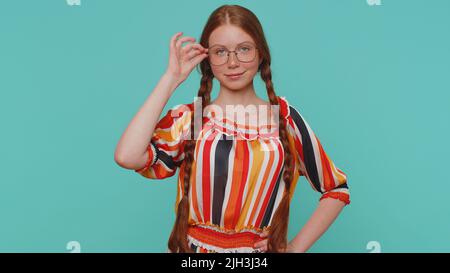 Fröhliche schöne rothaarige Mädchen in bunten Kleid lächelnd, Blick auf die Kamera. Junge Mode Modell Teenager Kind Kind trägt Brille Innenstudio Schuss isoliert auf blauem Hintergrund. Weibliche Natur Schönheit Stockfoto