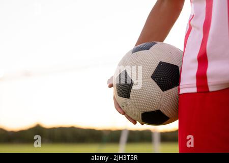 Mittelteil des männlichen kaukasischen Fußballspielers in rotem Jersey, der den Ball vor dem klaren Himmel hält Stockfoto