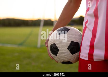 Mittelteil des männlichen kaukasischen Fußballspielers, der den Ball hält, während er bei Sonnenuntergang gegen den klaren Himmel steht Stockfoto