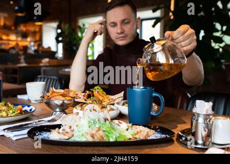 Ein Mann in einem Restaurant gießt sich eine Tasse heißen Tee aus einer Glas-Teekannen. Vorderansicht. Stockfoto