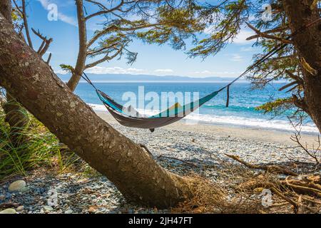 Romantische, gemütliche Hängematte im Schatten des Baumes am tropischen Strand am Meer. Perfekter paradiesischer Strand auf Vancouver Island mit Bäumen und Hängematte. Stockfoto