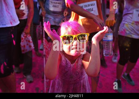Das süße kleine kaukasische Mädchen, das mit rosa Puder bedeckt ist und gelbe Töne trägt, hält beim Color Walk in Dubai, Vereinigte Arabische Emirate, die Hände mit einem Lächeln hoch Stockfoto