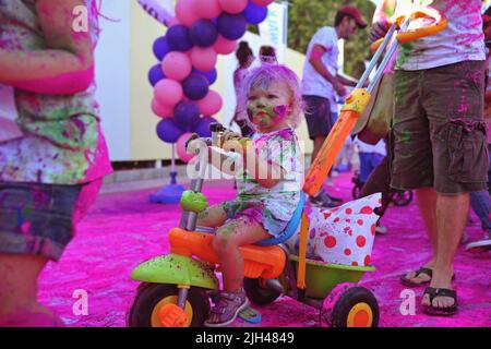 Niedliches kleines Mädchen, das mit buntem Puder im Push-Along-Dreirad für Kleinkinder bedeckt ist, sieht beim Color Walk in Dubai, Vereinigte Arabische Emirate, neugierig um sie herum aus. Stockfoto