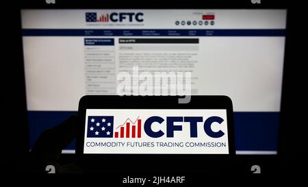 Person, die Mobiltelefon mit dem Logo der American Commodity Futures Trading Commission (CFTC) auf dem Bildschirm vor der Webseite hält. Konzentrieren Sie sich auf die Telefonanzeige. Stockfoto