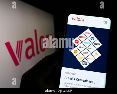 Person, die Mobiltelefon mit der Webseite der Schweizer Handelsgesellschaft Valora Holding AG auf dem Bildschirm vor dem Logo hält. Konzentrieren Sie sich auf die Mitte des Telefondisplays. Stockfoto