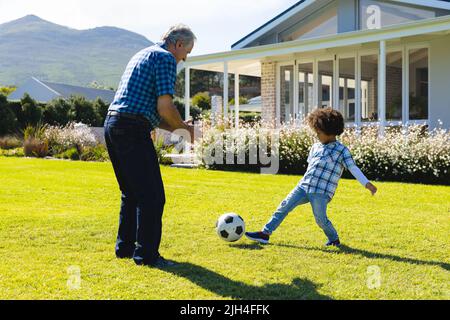 Kaukasischer Großvater spielt Fußball mit Biracial Enkel auf grasbewachsenen Land im Hof an sonnigen Tag Stockfoto