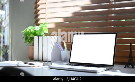 Computer-Laptop, Bücher, Kaffeetasse und Zimmerpflanze neben dem Fenster mit Sonnenlicht scheinen durch den Vorhang Stockfoto