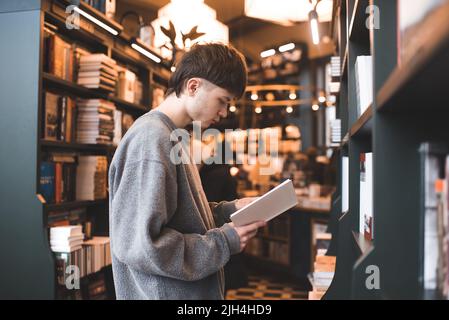 Stilvoll schöner Teenager Junge Student 18-19 Jahre alt lesen Papierbuch in der öffentlichen Bibliothek oder Buchhandlung innen. Millennials Lifestyle. Zurück zur Schule. Stockfoto