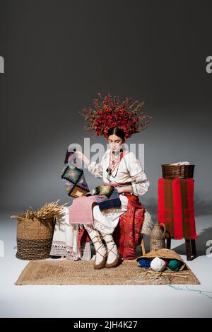 Die ganze Länge der brünetten ukrainischen Frau in traditioneller Kleidung und roten Blumenkranz mit gestricktem Schal auf dunkelgrau Stockfoto