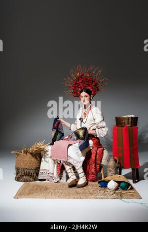 Die ganze Länge der jungen ukrainischen Frau in traditioneller Kleidung und Blumenkranz mit Beeren hält gestrickten Schal auf dunkelgrau Stockfoto