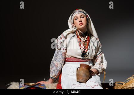 Junge ukrainische Frau in traditioneller Kleidung mit Ornament, das Tontopf hält und auf einer schwarzen Bank sitzt Stockfoto