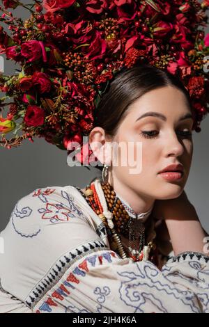 Porträt einer hübschen ukrainischen Frau in traditioneller Kleidung und Blumenkranz mit roten Beeren isoliert auf grau Stockfoto