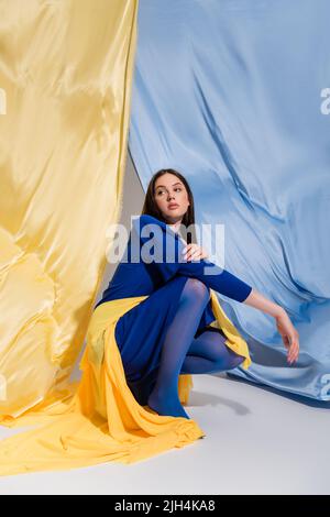 Trendige junge ukrainische Frau in stilvoller Blockkleidung sitzt in der Nähe der blauen und gelben Flagge Stockfoto