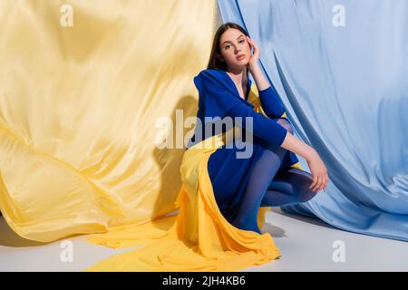Junge ukrainische Frau in Blockkleidung, die in der Nähe von blauem und gelbem Stoff sitzt Stockfoto