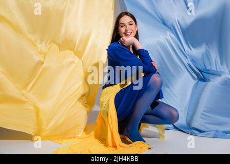 Glückliche ukrainische Frau in stilvoller Blockkleidung, die in der Nähe von blauem und gelbem Stoff sitzt Stockfoto