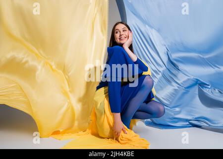 Fröhliche ukrainische Frau in stilvoller Blockkleidung, die in der Nähe von blauem und gelbem Stoff sitzt Stockfoto