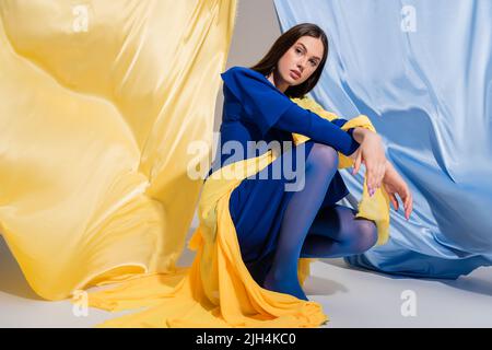 Junge ukrainische Frau in stilvoller Blockkleidung, die in der Nähe von blauem und gelbem Stoff sitzt Stockfoto