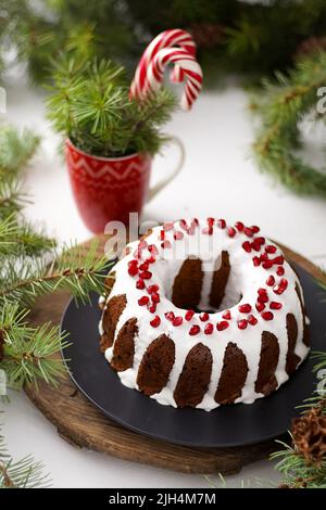 Weihnachtliches Essen. Runde Torte mit weißer Glasur. Cupcake mit einem Loch in der Mitte mit Granatapfel. Grüne Fichtenzweige auf dem Tisch. Stillleben. Süßes Gebäck, Desserts und Leckereien für das neue Jahr. Stockfoto