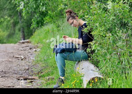 Junge Frau sitzt auf einem Baumstamm am Rande eines Waldwegs und sucht etwas in ihrer Tasche, Seitenansicht, Deutschland, Nordrhein-Westfalen, Stockfoto