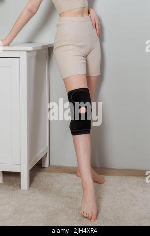 Schwarze Kniestütze auf einem Frauenbein. Mädchen in einer Orthese im Inneren des Hauses. Orthopädische anatomische Hosenträger zur Kniefixierung, Verletzungen und Stockfoto