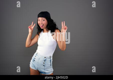 Junges chinesisches Mädchen mit Bandana und Shorts, das Siegeszeichen macht, in einer lustigen Pose und Ausdruck Stockfoto