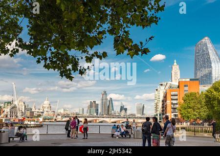 London, England, UK-Juli 21 2019: Junge Menschen versammeln sich im Schatten der Bäume, entlang der Uferpromenade und bewundern die ikonische Skyline von London City auf der anderen Seite Stockfoto