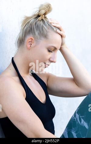 Kaukasische blonde junge Sportlerin mit der Hand auf dem Kopf, die Kopfschmerzen und Depressionen durch Sportverletzungen bekommt. Verliererin Frau ist erschöpft und depressiv und sitzt traurig auf der Straße. Hochwertige Fotos Stockfoto