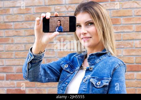 Attraktive blonde kaukasische Influencerin Frau macht Selbstporträt auf Smartphone-Bildschirm über rotem Backstein Hintergrund. Hochwertige Fotos Stockfoto