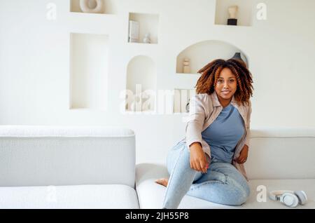 afroamerikanische Millennial-Mädchen, zufrieden und glücklich, sitzen auf einem bequemen Sofa in gemütlichen modernen hellen Wohnzimmer. Genießen Sie ihr Leben Stockfoto