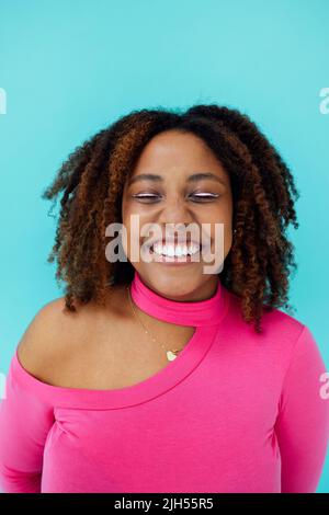 Verträumte junge, schöne afroamerikanische Frau mit hellen Augenlinien, die einen pinken Body über der blauen Wand trägt, hält ihre Augen geschlossen, sieht mit glücklichen Ausblicken aus Stockfoto