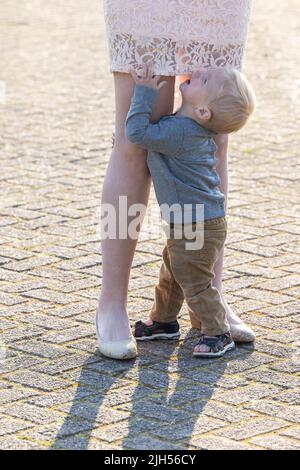 Trauriger kaukasischer blonder kleiner Junge, der ihre Mutter an den Beinen umarmt. Hochwertige Fotos Stockfoto