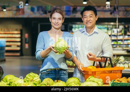 Junges Ehepaar, asiatischer Mann und blonde Frau im Supermarkt, Käufer wählen Gemüse im Gemüsebereich, Porträt von glücklichen Käufern, lächelnd Stockfoto