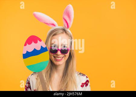 Festliche kaukasische christliche Mädchen in Sonnenbrillen und weißen Hasenohren halten Karton Osterei über orangenen Hintergrund. Studio-Porträt. Speicherplatz kopieren. Hochwertige Fotos Stockfoto