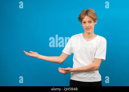 Der junge Teenager zeigt die Handflächen isoliert auf blau zur Seite. Freundlicher, gut aussehender Junge, der seine Aufmerksamkeit auf den Kopierbereich, das beste Angebot, den Verkauf und die Präsentation von Neuheit lenkt Stockfoto